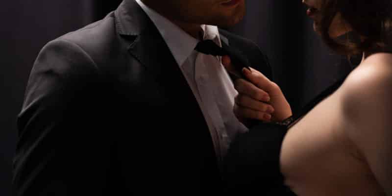 cropped view of seductive woman pulling tie of man in suit on black