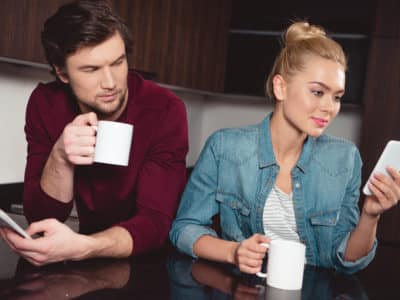 jealous husband holding coffee cup and looking at wife using smartphone in kitchen