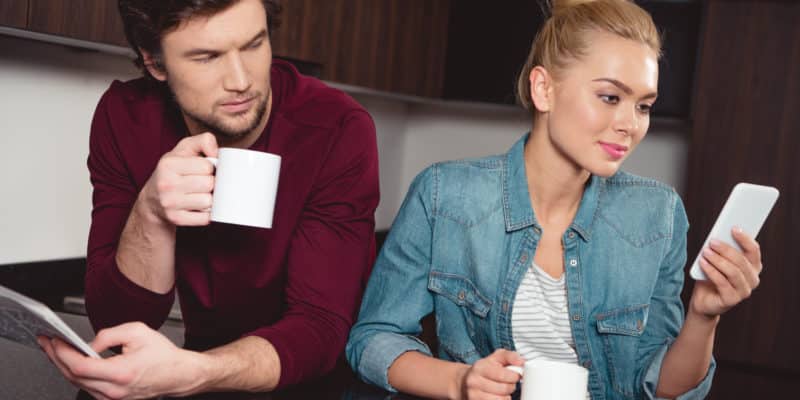 jealous husband holding coffee cup and looking at wife using smartphone in kitchen